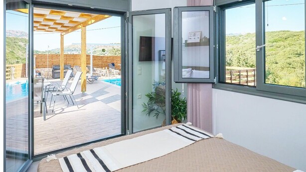 A bedroom with a bed, a TV, a sliding glass door to a balcony with a pool, and a view of the hills. - Villa Eleonas with private swimming pool (Rethymno)