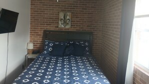 A bedroom with a brick wall, a ceiling fan, a bed with patterned bedding, a nightstand with a lamp, and a flat-screen TV mounted on the wall.
