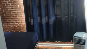 A room with a brick wall, a bed, a window with dark curtains, and an air conditioning unit.