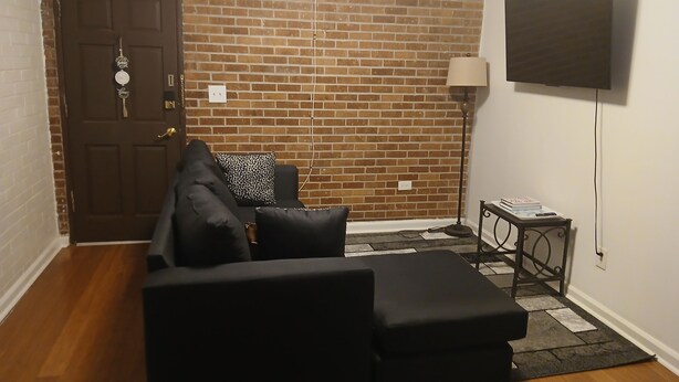 A room with a brick wall, a dark brown door, a black sofa, a patterned throw pillow, a small side table with a lamp, and a flat-screen TV mounted on the wall.
