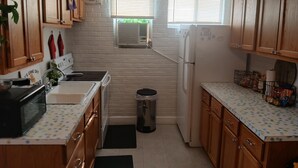 A compact kitchen with wooden cabinets, a microwave, a refrigerator, and a sink. There are two windows with blinds, a wall-mounted fan, and a trash bin.