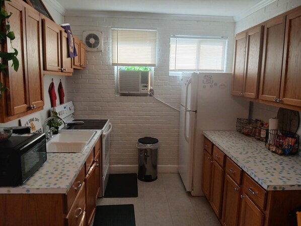 A compact kitchen with wooden cabinets, a microwave, a refrigerator, and a sink. There are two windows with blinds, a wall-mounted fan, and a trash bin.