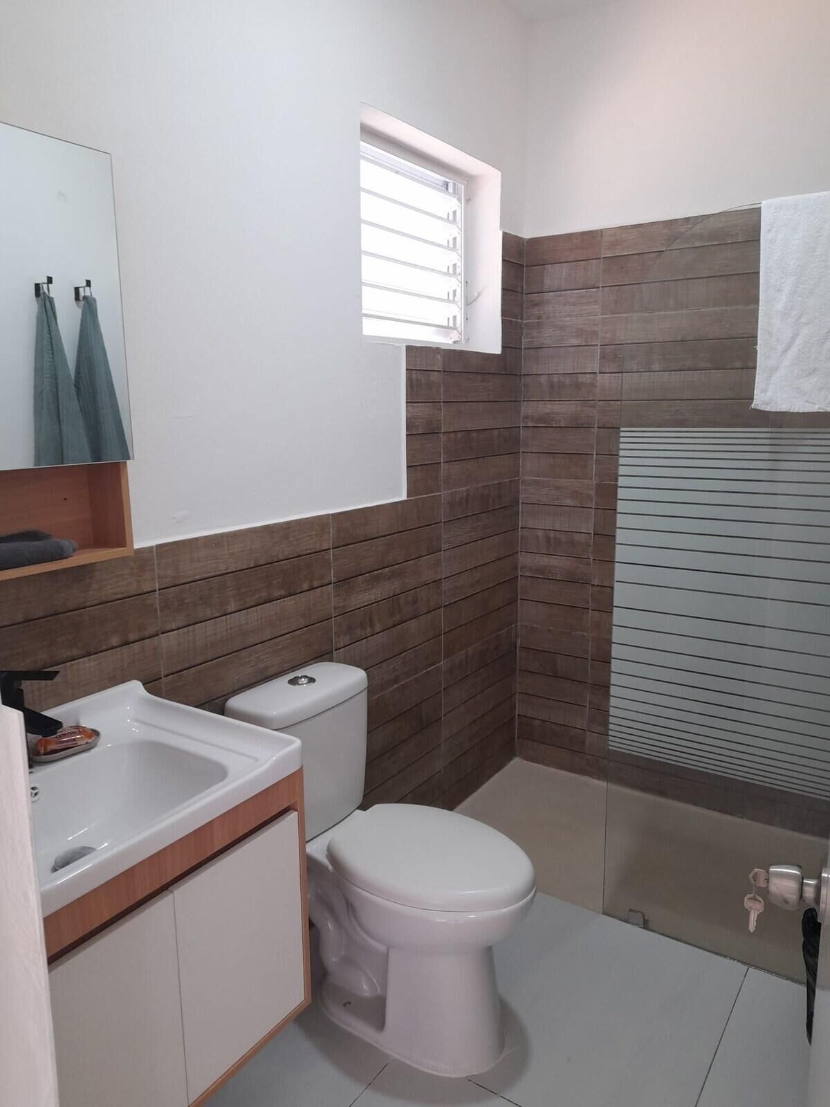 A small bathroom with a toilet, sink, and mirror. There is a window above the toilet and a towel hanging on the wall.