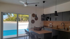 A modern kitchen with a central island, dark cabinetry, pendant lighting, and a view of a swimming pool through glass doors. - Villa Sleeps 8 With Private Pool (Sosúa)