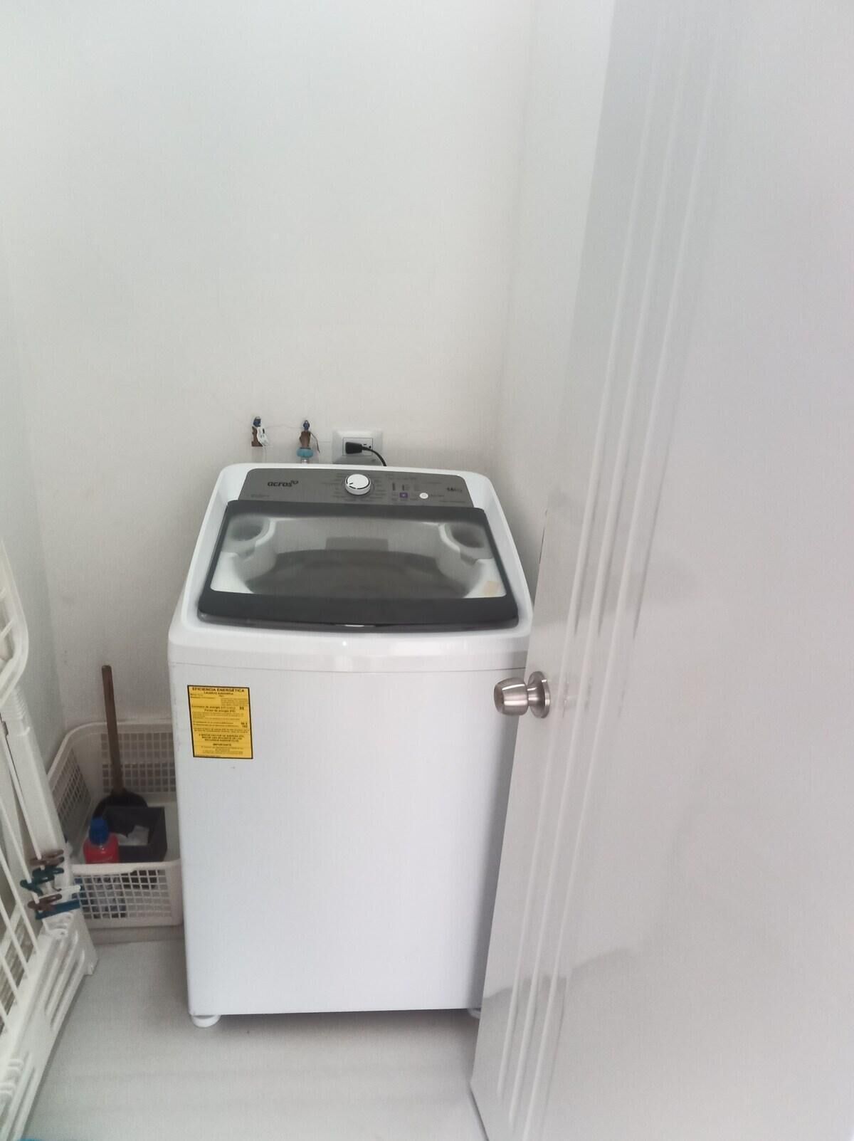 A compact, white washing machine in a small, enclosed space with a door, a broom, and a laundry basket.
