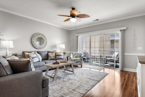 Un salon moderne avec un ensemble de canapés gris, une table basse et un fauteuil à motifs. On y trouve un ventilateur de plafond, un grand miroir et, à travers une porte-fenêtre coulissante, une vue sur un patio extérieur.