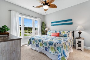 A bedroom with a large bed, a ceiling fan, a window with white curtains, a bedside table with a lamp, and a view of the outdoors.