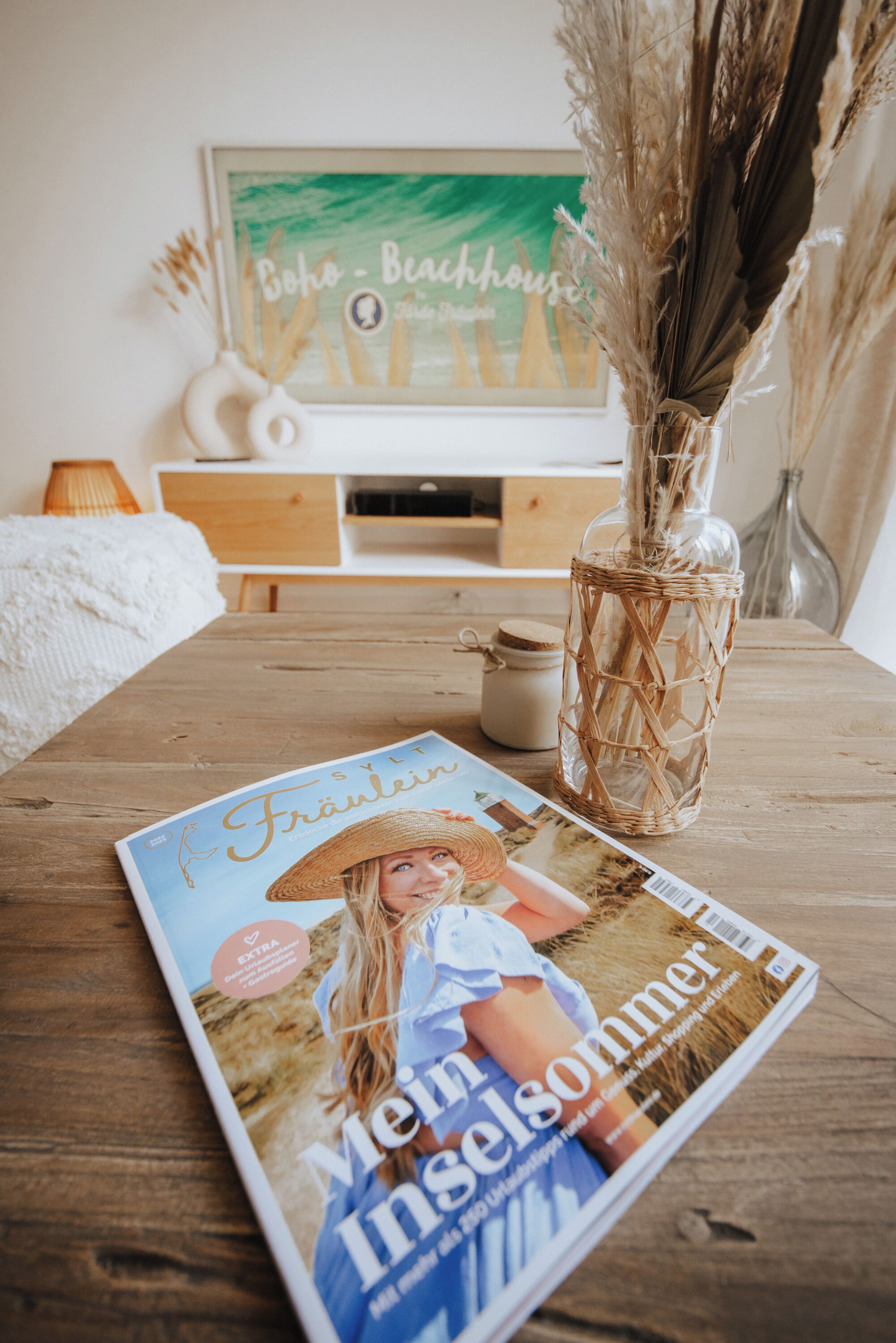 Un magazine sur une table en bois, un vase en osier contenant des plantes séchées et un tableau encadré accroché au mur.