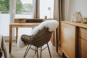 A room with a wooden desk, a wicker chair with a white fur throw, a wooden dresser, and a window with curtains.