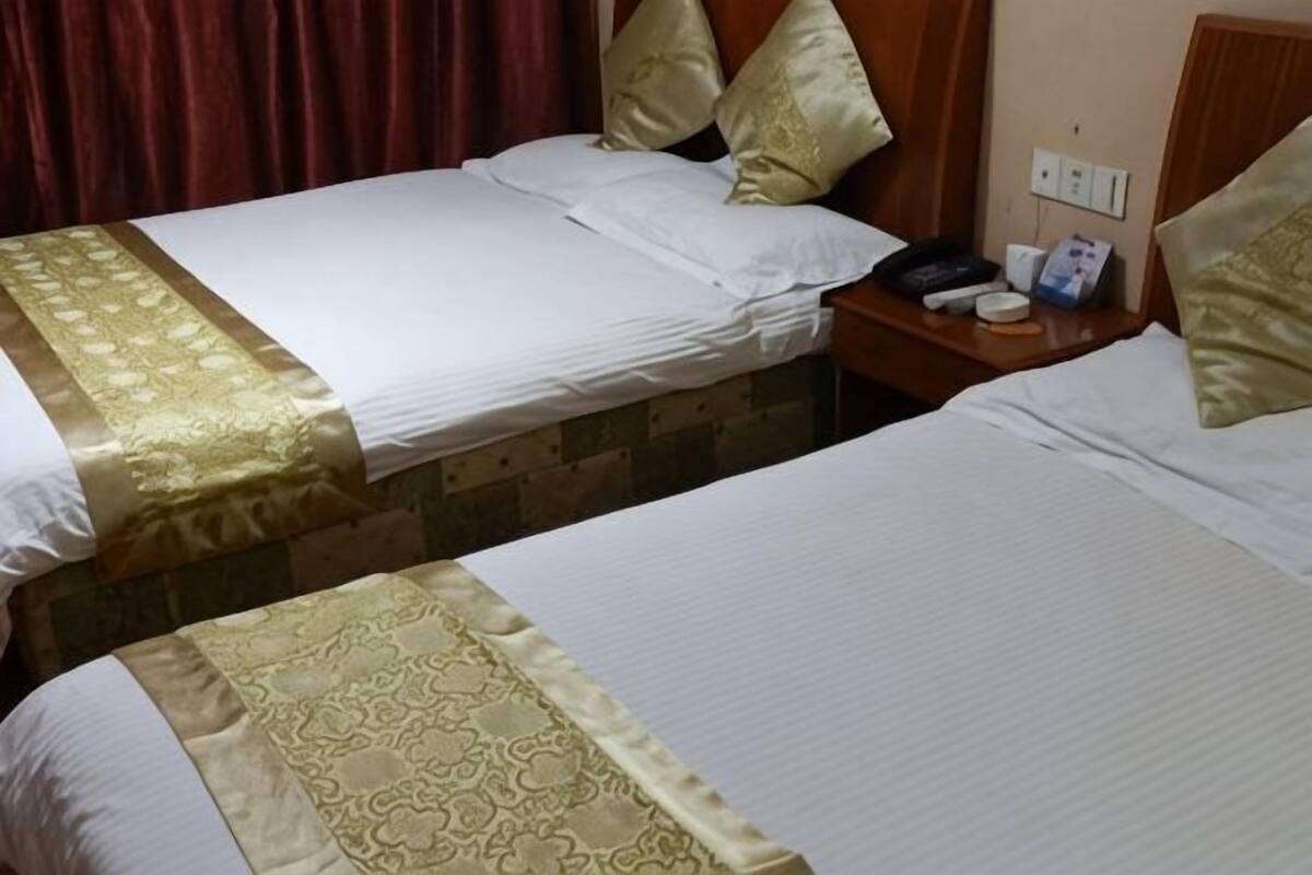 Dos camas individuales con ropa de cama dorada y blanca, una mesita de noche de madera con un teléfono y un objeto pequeño, y una cortina roja.