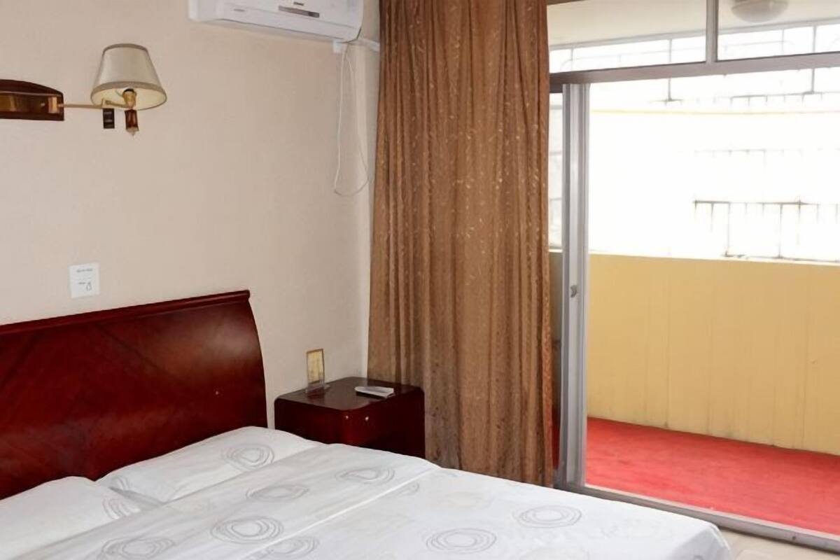 Habitación de hotel con cama, mesita de noche, lámpara y vistas a un balcón.