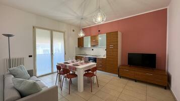 Un soggiorno moderno con un divano, un tavolo da pranzo per quattro persone, un angolo cottura con pensili e una TV, e un'ampia finestra con vista.