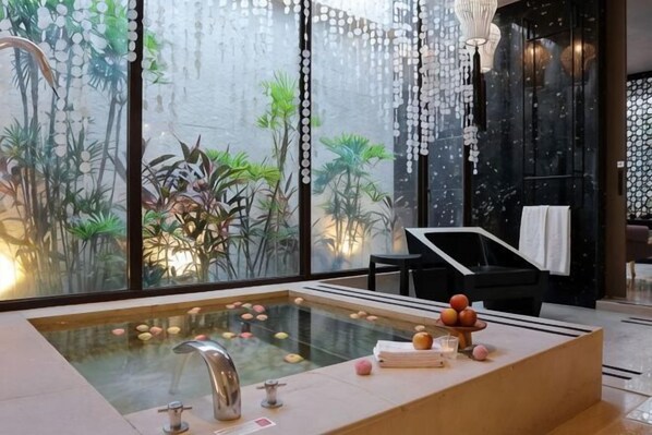 A modern spa room with a large bathtub, a black sofa, and a view of indoor plants through large windows.
