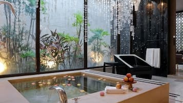 Uma área de spa moderna com uma grande banheira de hidromassagem envidraçada, uma poltrona de descanso preta e vista para plantas em interior.