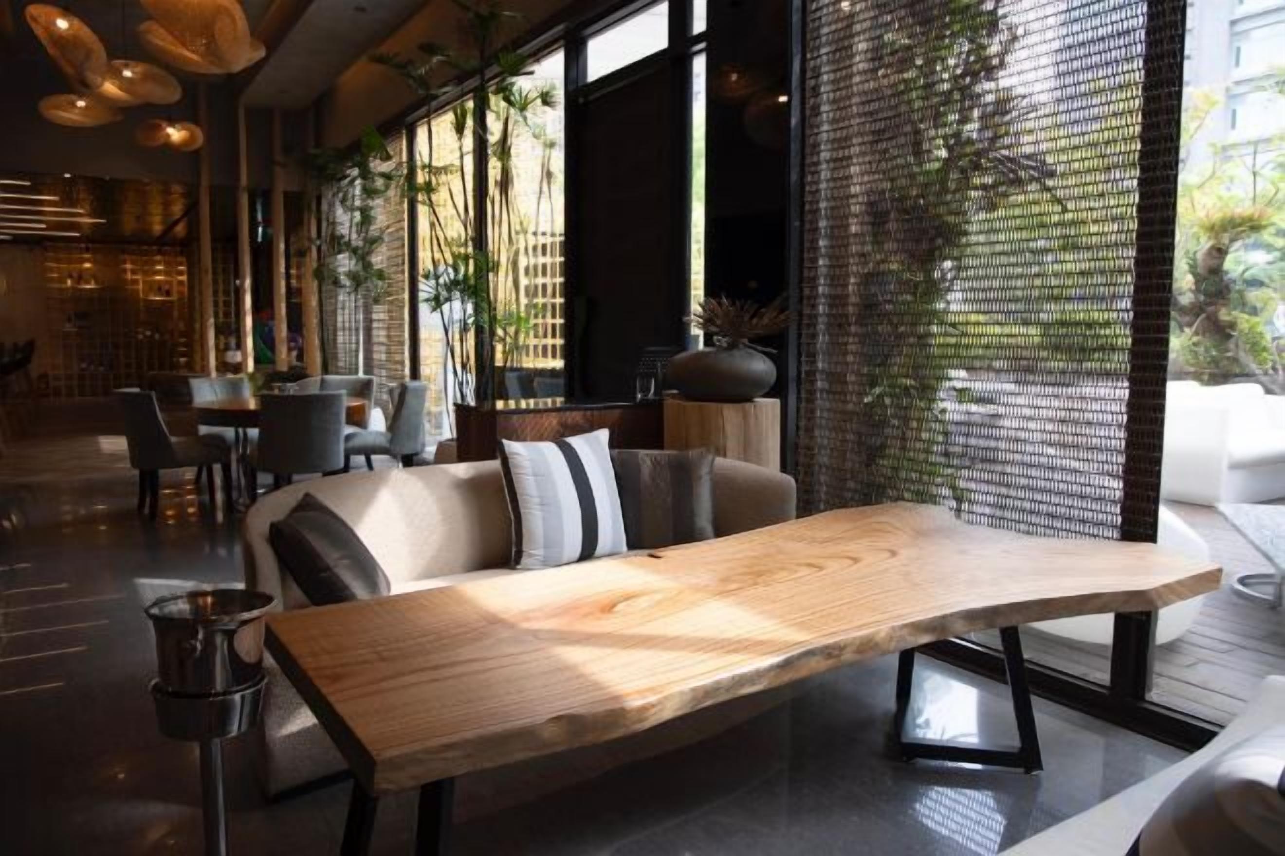 Um interior moderno com uma mesa de madeira comprida, um sofá com almofadas e vista para uma parede de tijolos através de uma janela.
