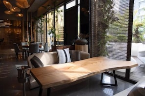 Um interior moderno com uma mesa de madeira comprida, um sofá com almofadas e vista para uma parede de tijolos através de uma janela.