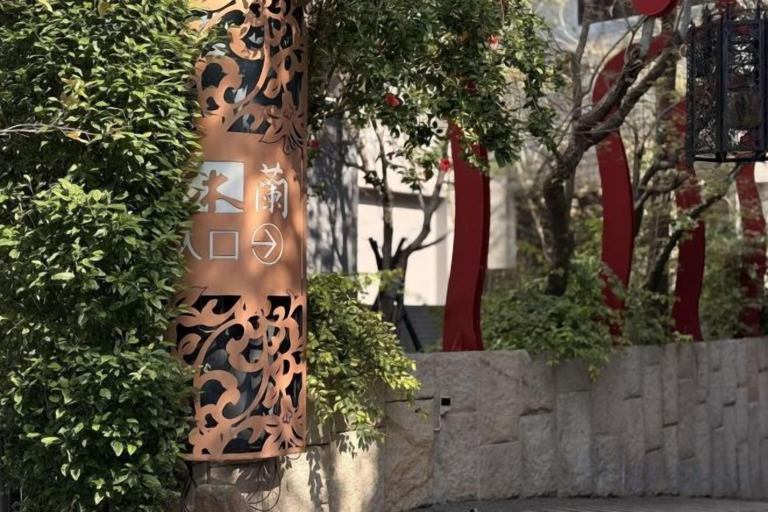 A decorative metal structure with intricate cut-out patterns, situated in a landscaped area with a stone wall and greenery.