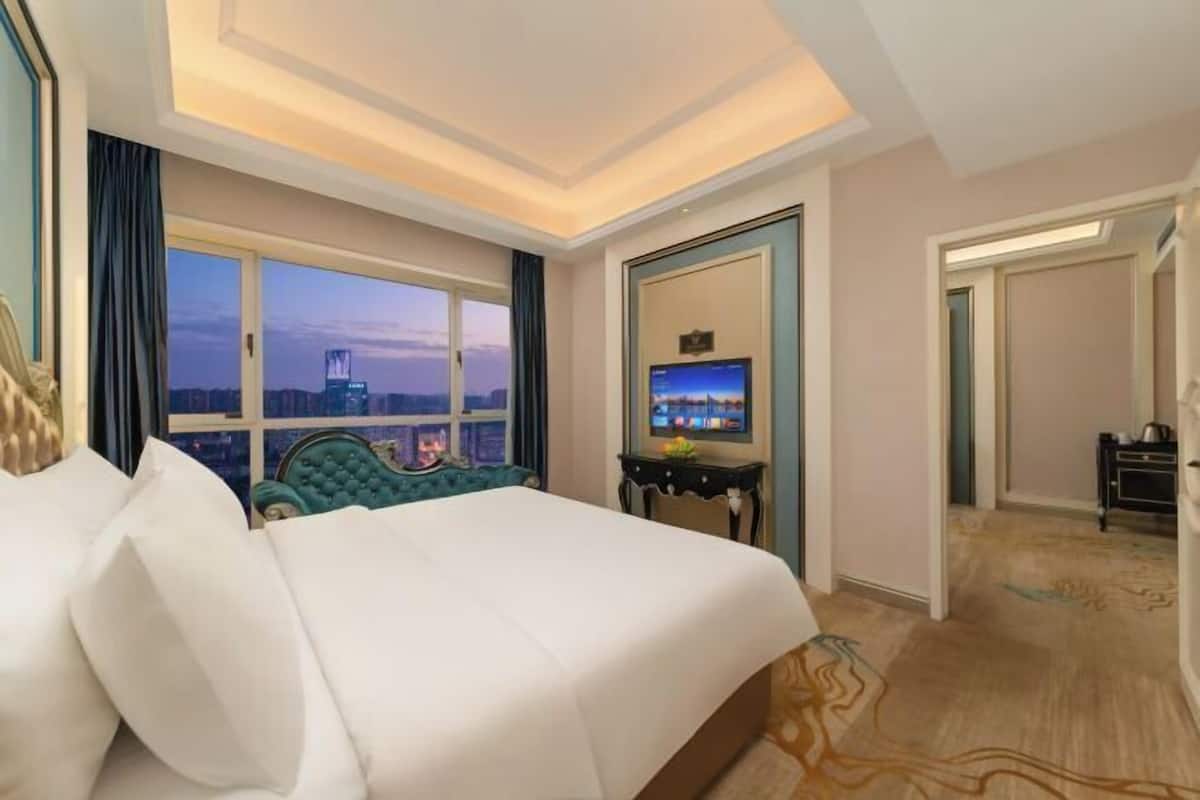 A hotel room with a large bed, a television on a stand, a window with a city view, and a door leading to another room.