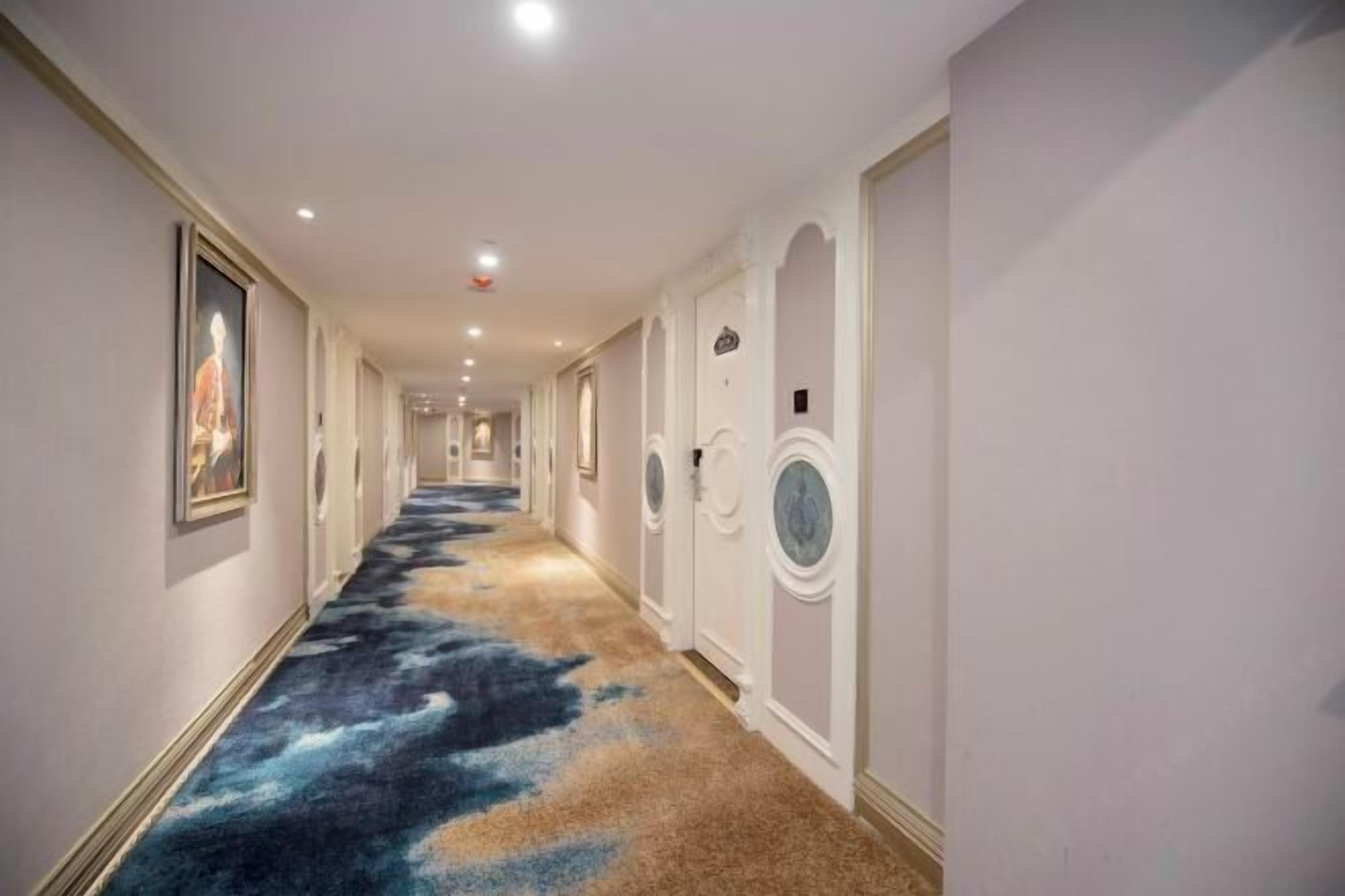 A hotel corridor with a patterned carpet, white walls, and doors with circular windows.