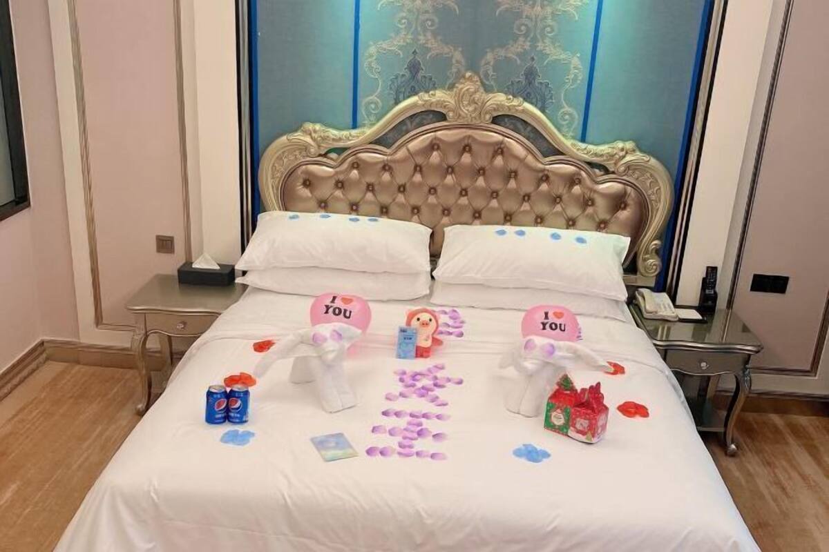 A hotel room with a large bed, two pillows, a decorative headboard, and a bedside table with a lamp.