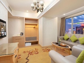 A hotel room with a seating area, a small table, a sofa with green cushions, a wooden cabinet, and a large window with a city view. - Vienna International Hotel (Changsha)