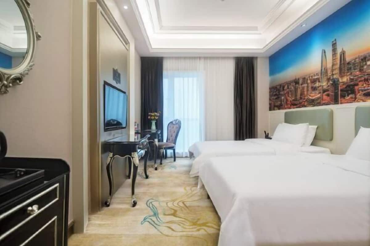 A hotel room with two beds, a desk, a chair, and a large painting of a cityscape.
