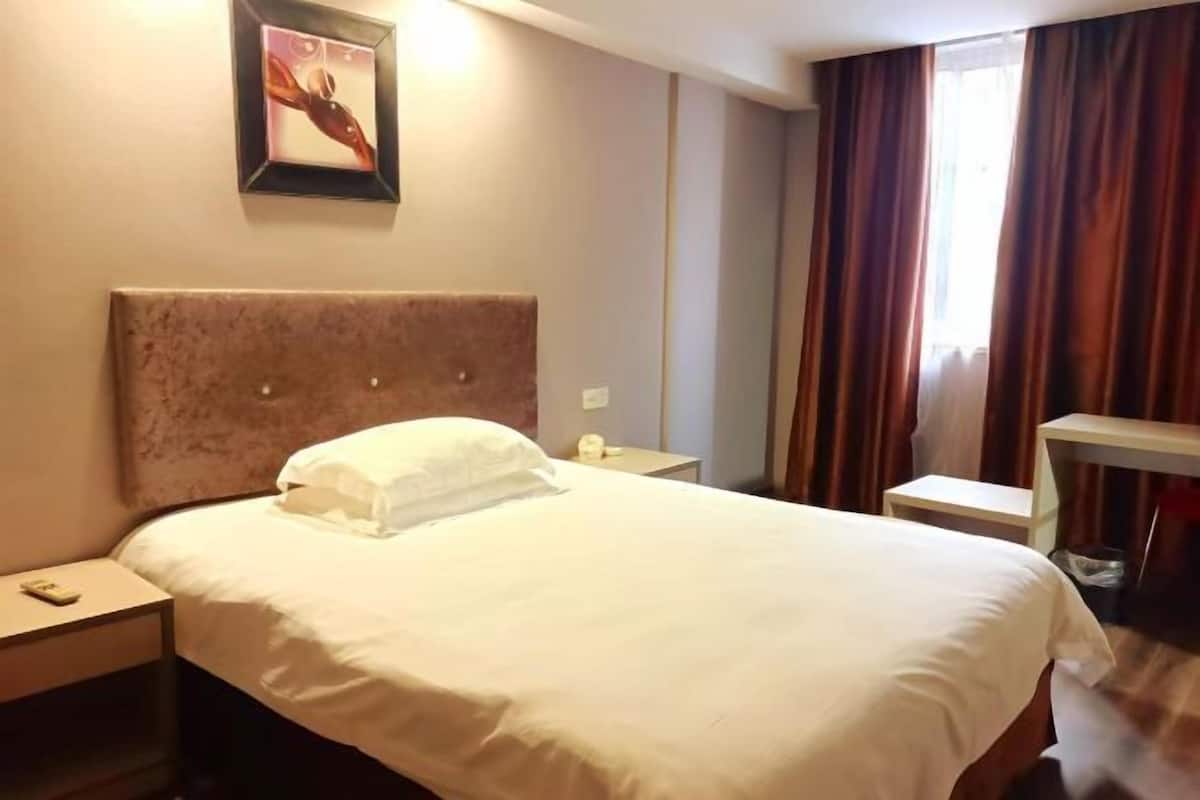 A hotel room with a large bed, a headboard, a bedside table with a lamp, a framed picture on the wall, and a window with curtains.