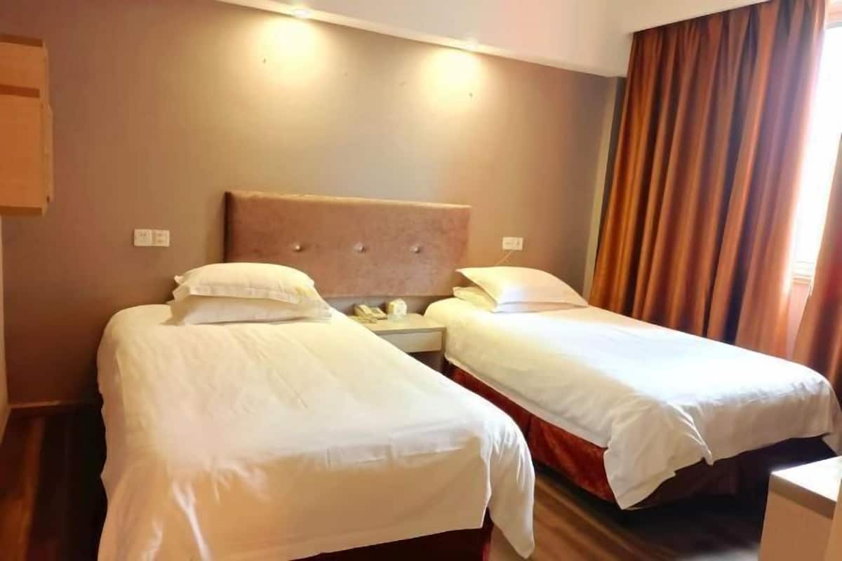 A hotel room with two single beds, a wooden headboard, a nightstand, and a window with curtains.