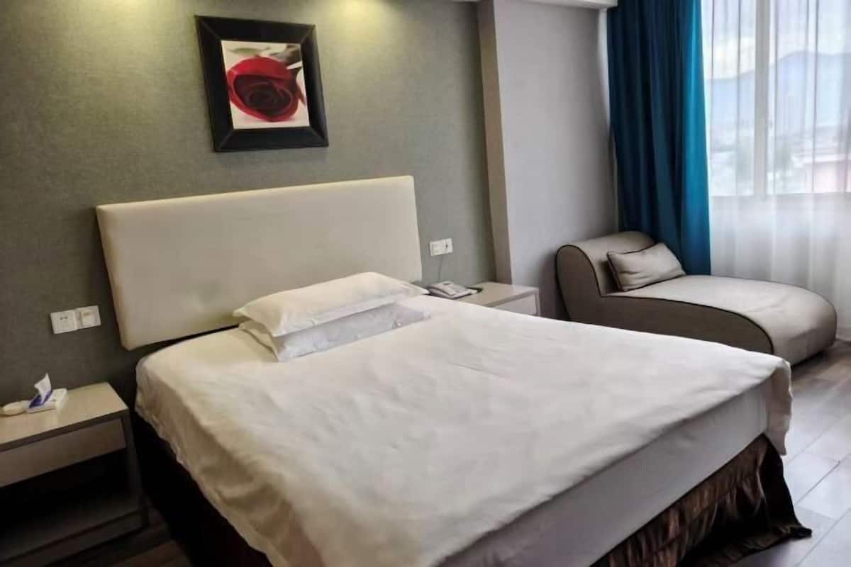 A hotel room with a large bed, a bedside table, a chair, a framed picture on the wall, and a window with curtains.