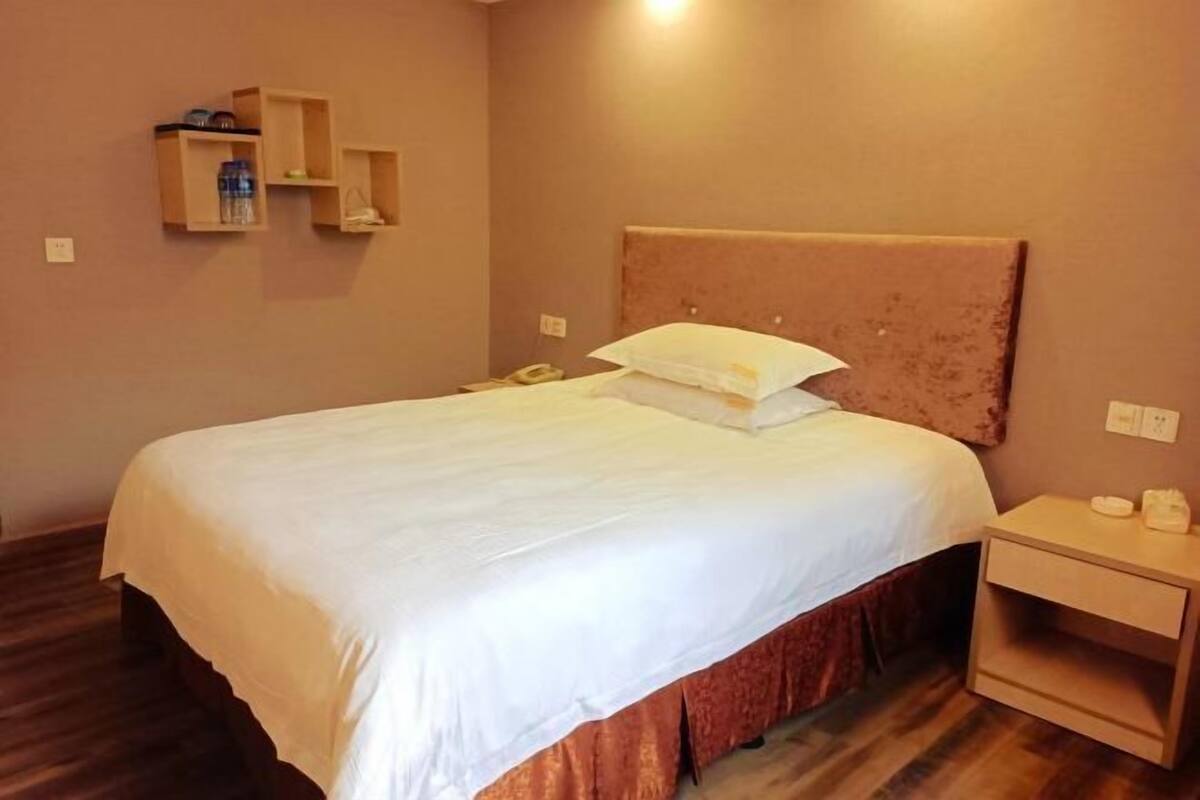 A hotel room with a bed, a nightstand, and a shelf with a bottle and a phone.