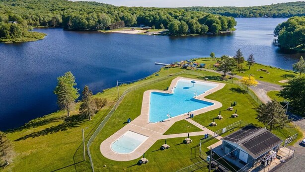 Une grande piscine de forme irrégulière, entourée d’une pelouse bien entretenue, d’où l’on peut admirer un lac et des collines boisées en arrière-plan.