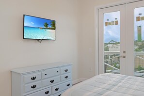 Ein Schlafzimmer mit einem Bett, einer weißen Kommode mit Schubladen, einem Fernseher, der eine Strandansicht zeigt, und einem Balkon mit Geländer.
