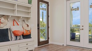 A room with a wooden floor, a white built-in bench with bags and pillows, a brown door, and a glass door with a view of the outside. - It's A Wonderful Life (Port St. Joe)