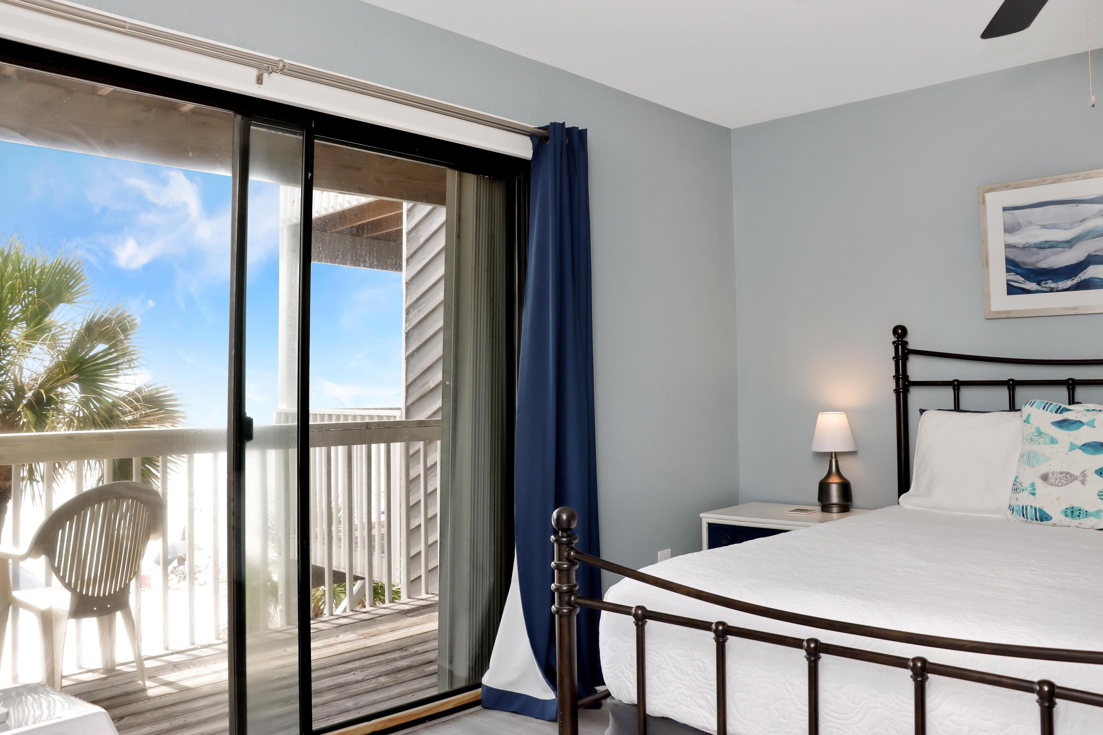 A bedroom with a bed, a nightstand, a lamp, and a view of a balcony with a chair.