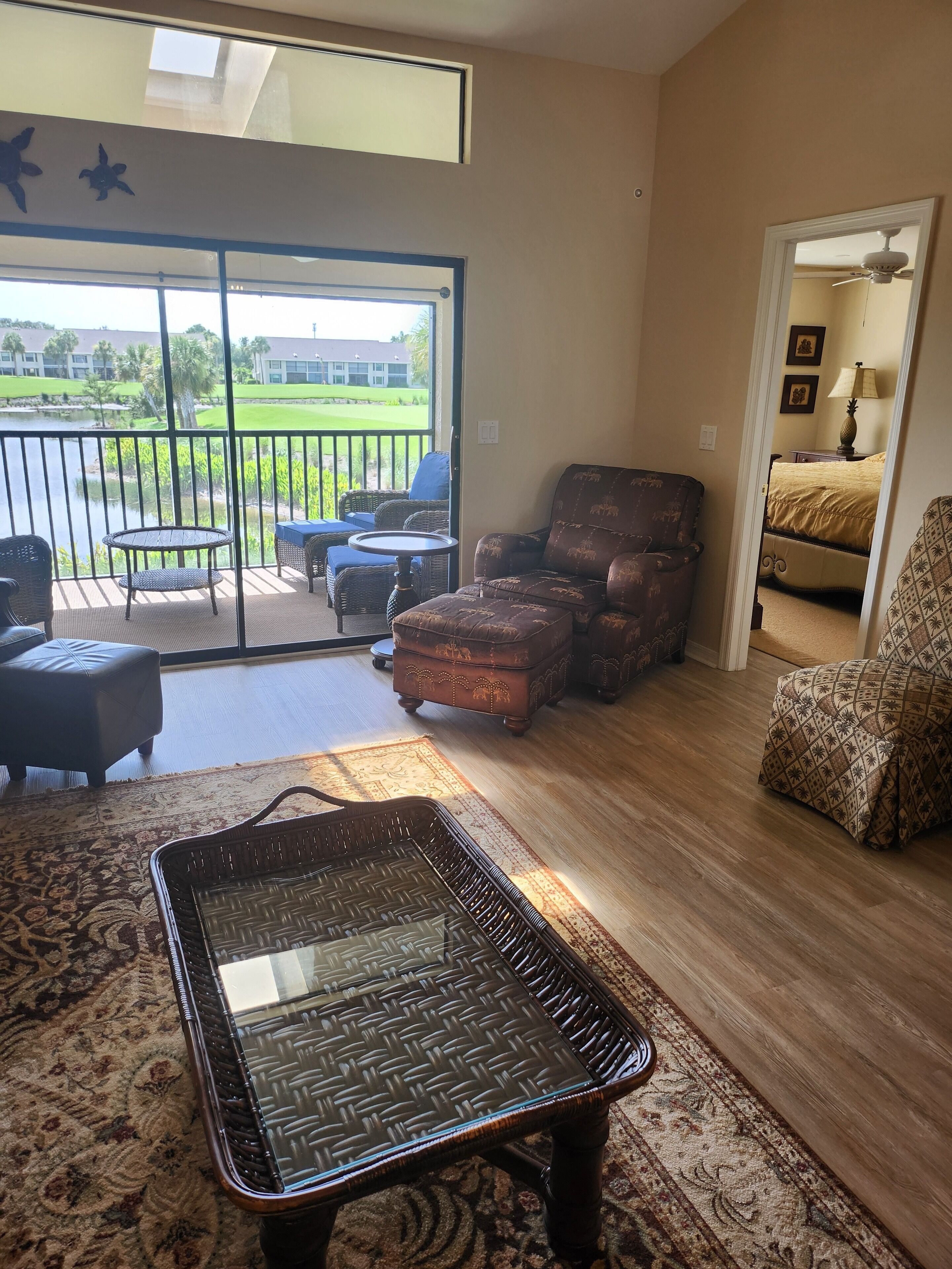 A spacious living area with a large sliding glass door leading to a balcony with a view of a golf course. There is a wicker chair, a small round table, and a patterned rug on the floor.