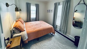 A bedroom with a bed, a bedside table, a lamp, a mirror, and a window with curtains.