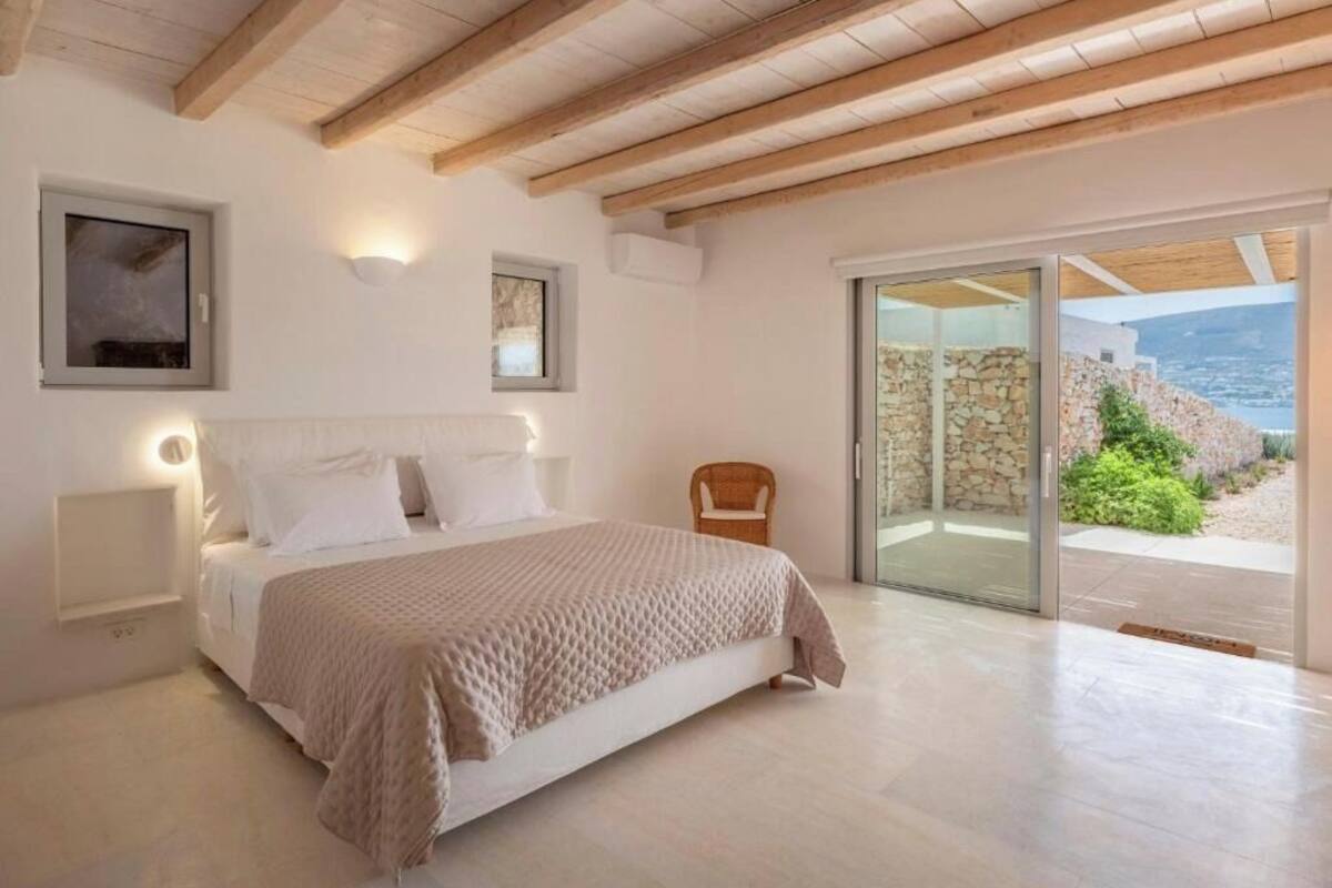 Quarto com cama grande, vigas de madeira, porta de correr de vidro para área externa e parede de pedra.
