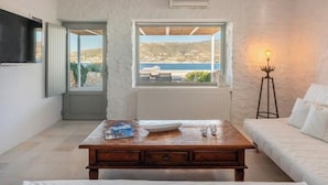 A room with a wooden table, white sofa, and a view of the sea through a window. - Donum Dei Seaside Suites & Villa (Paros)