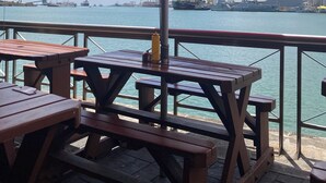 A waterfront patio with wooden tables and benches, an umbrella, and a view of the harbor with ships and cranes. - Space Villa 3 (Grand Baie)