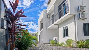 A modern building with a gravel driveway, air conditioning units, and a small garden area with plants and a decorative stone feature. - Space Villa 3 (Grand Baie)