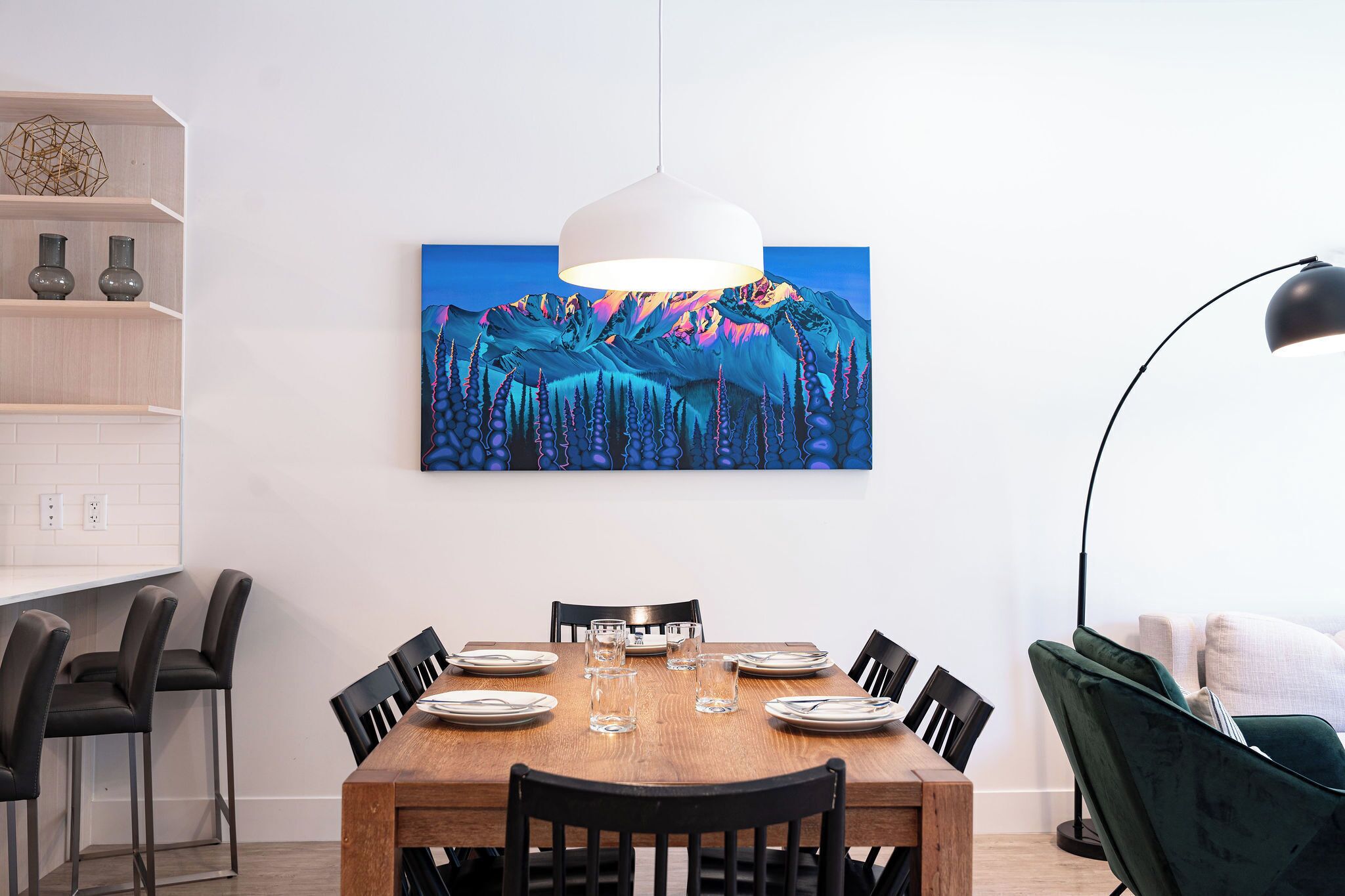 A dining area with a wooden table set for six, black chairs, a white pendant light, and a painting of a mountain landscape on the wall.