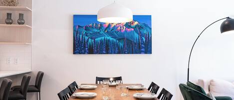 A dining area with a wooden table set for six, black chairs, a white pendant light, and a painting of a mountain landscape on the wall.