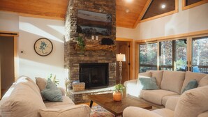 A cozy living room with a stone fireplace, beige sofas, and large windows offering a view of the outdoors. - McStay in McCay - Charming 4-bed cabin w/ outdoor fireplace and large jacuzzi!  (McCaysville)