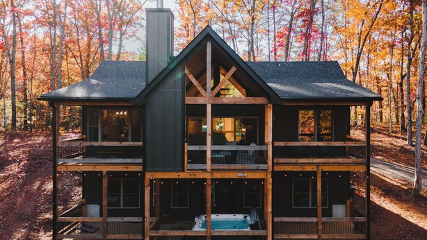 A two-story log cabin with a central chimney, multiple windows, and a deck area. - McStay in McCay - Charming 4-bed cabin w/ outdoor fireplace and large jacuzzi!  (McCaysville)