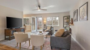 A spacious living room with a flat-screen TV, a ceiling fan, and a large window providing a view of the outdoors. - Set & Drift | Hatcher Pass, Pets Allowed, Peaceful (Palmer)
