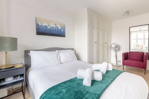 A hotel room with a bed, a desk with a lamp, a chair, a painting, and a window.