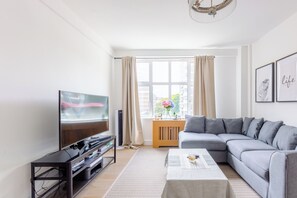 A modern living room with a flat-screen TV, grey sofa, wooden cabinet, and a large window with curtains.