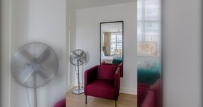 Ein Zimmer mit einem Standventilator, einem roten Sessel, einem Spiegel und einem Fenster mit Blick auf Gebäude.