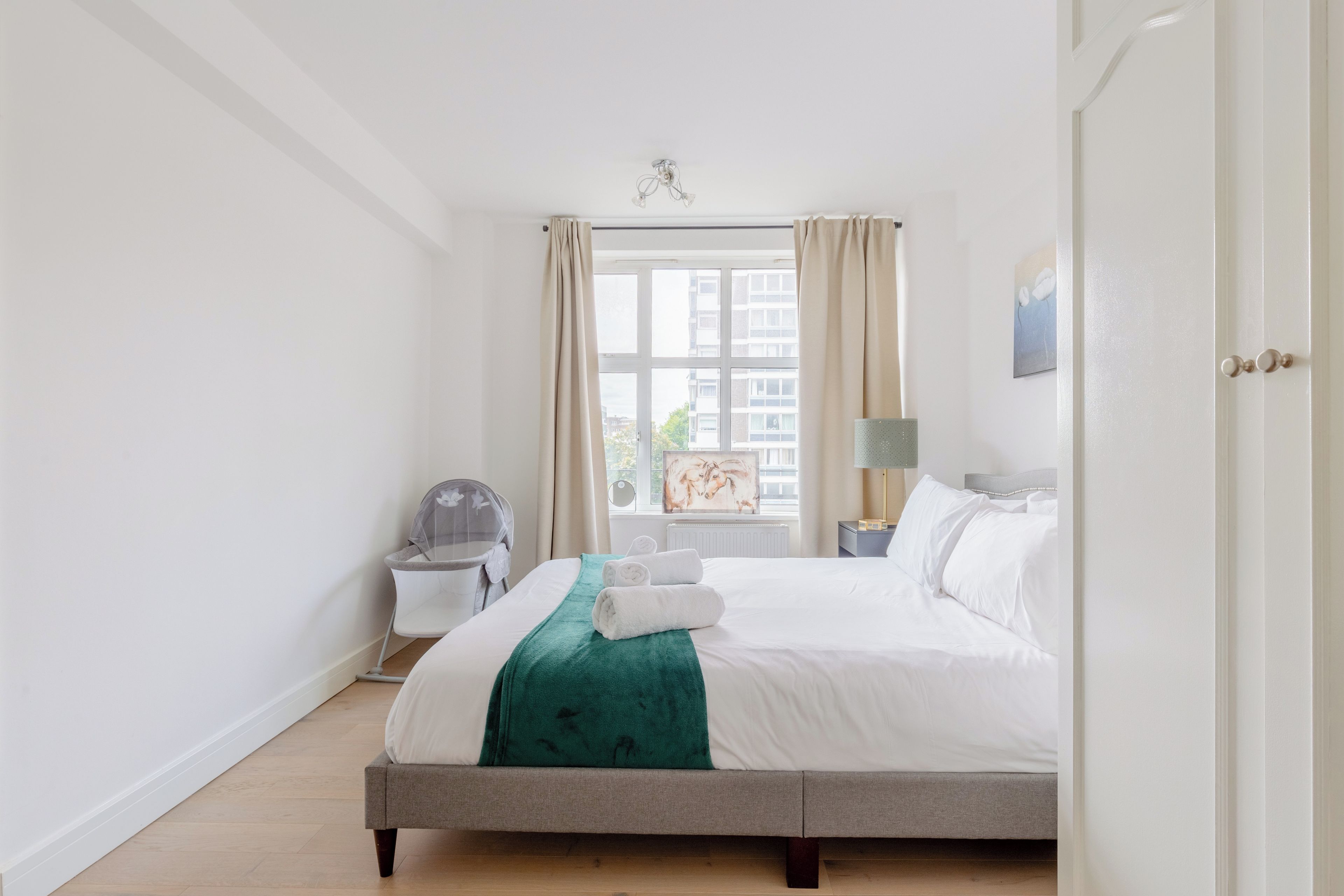A hotel room with a bed, a window with curtains, a bedside table, a lamp, and a painting on the wall.