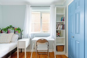 Una habitación con un escritorio blanco, una silla, una estantería, una ventana con cortinas y una puerta azul.